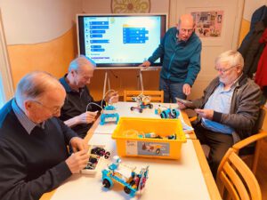 Vier Männer bauen Lego-Roboter, im Hintergrund ein Bildschirm mit Programmierschritten