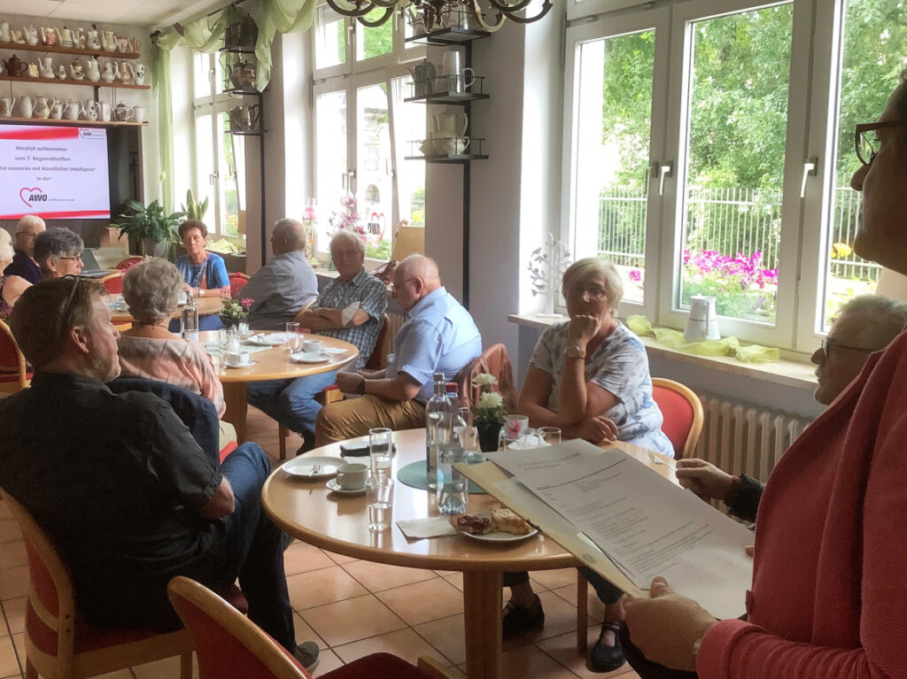 Mehrer ältere Menschen sitzen um Tische, im Hintergrund ein großer Bildschirm.