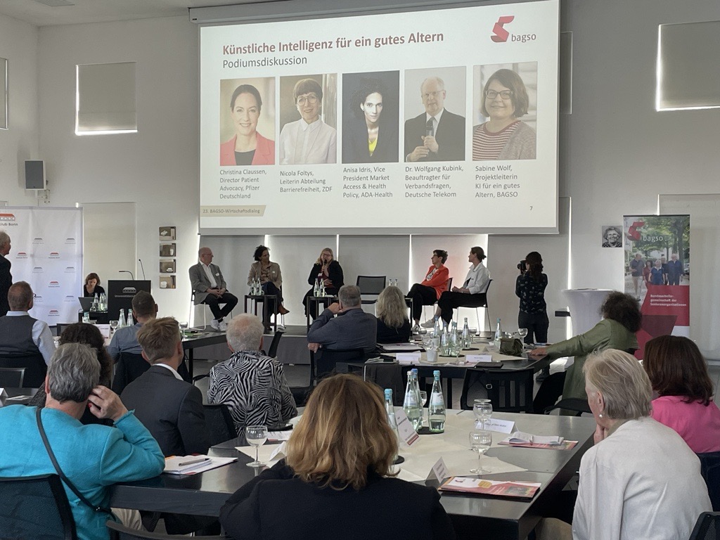 bei einer Veranstaltung mit viel Publikum findet auf der Bühne eine Podiumsdiskussion statt. Beteiligt sind fünf Menschen. Auf einer Leinwand über ihnen ist zu erkennen, dass sie über das Thema "KI für ein gutes Altern" diskutieren.