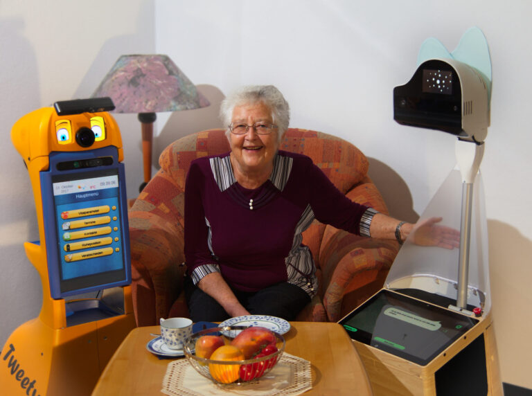 Eine ältere Frau sitzt lächelnd zwischen zwei Assistenzrobotern.