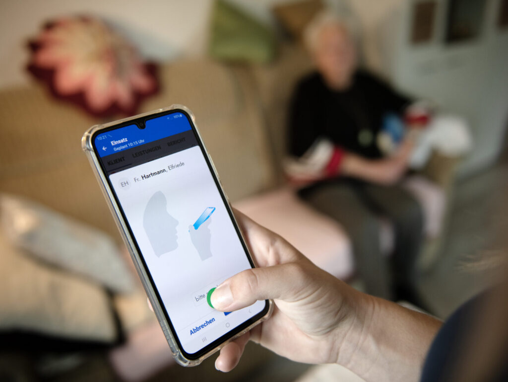 Smartphone mit digitaler Spracheingabe, im Hintergrund Person auf einem Sofa