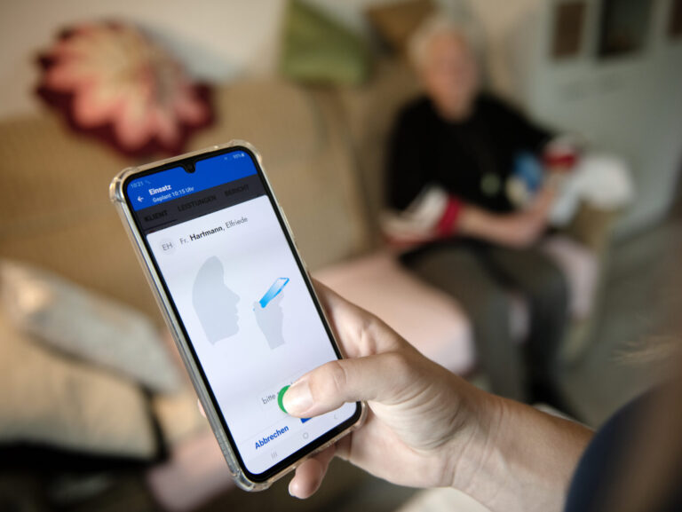 Smartphone mit digitaler Spracheingabe, im Hintergrund Person auf einem Sofa
