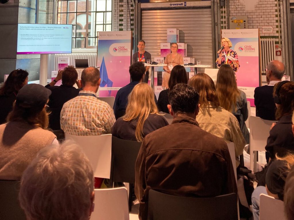 Podiumsdiskussion mit drei Personen, im Vordergrund Publikum, das aufmerksam zuhört