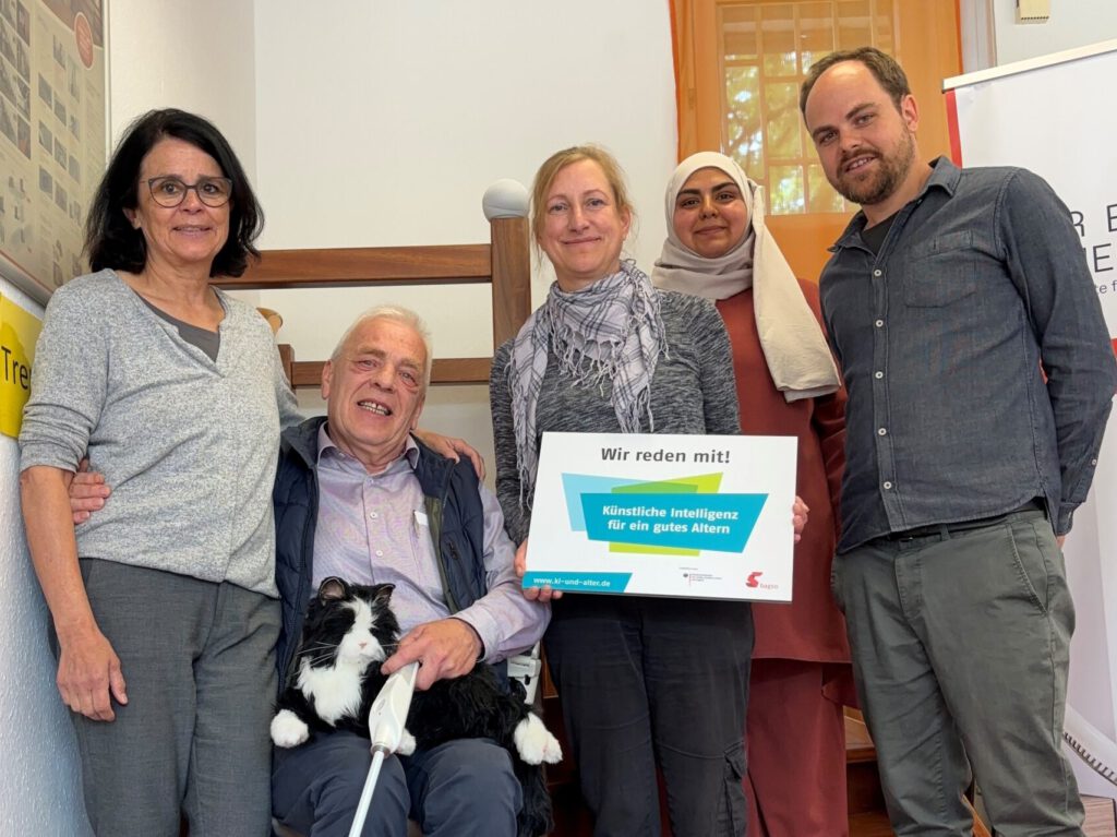 Fünf unterschiedliche Personen präsentieren ein Schild "Wir reden mit, KI für ein gutes Altern". Darunter ein älterer Mann, auf dessen Beinen eine Roboterkatze liegt.