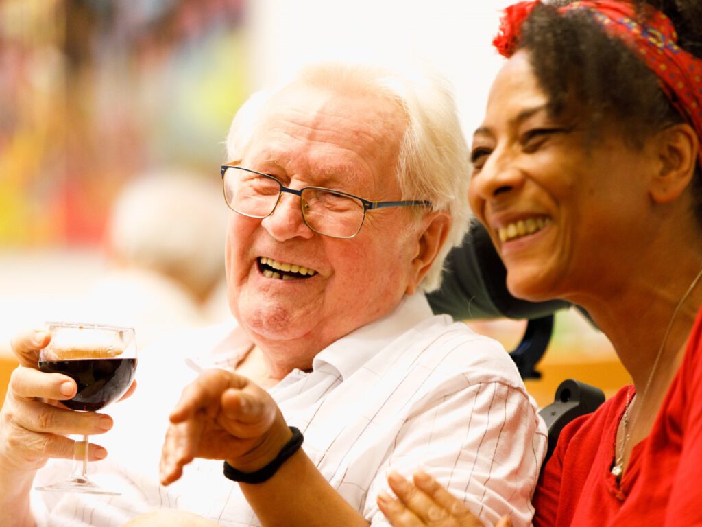 Ein älterer Mann trinkt ein Glas Wein. Eine jüngere Frau sitzt neben ihm. Beide lachen herzlich.