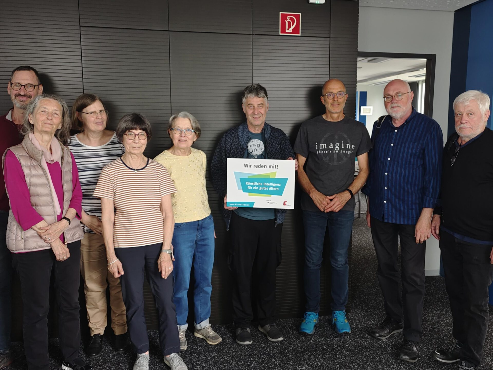 Eine Gruppe mit neun Personen präsentieren ein Schild auf dem steht "Wir reden mit. KI für ein gutes Altern"