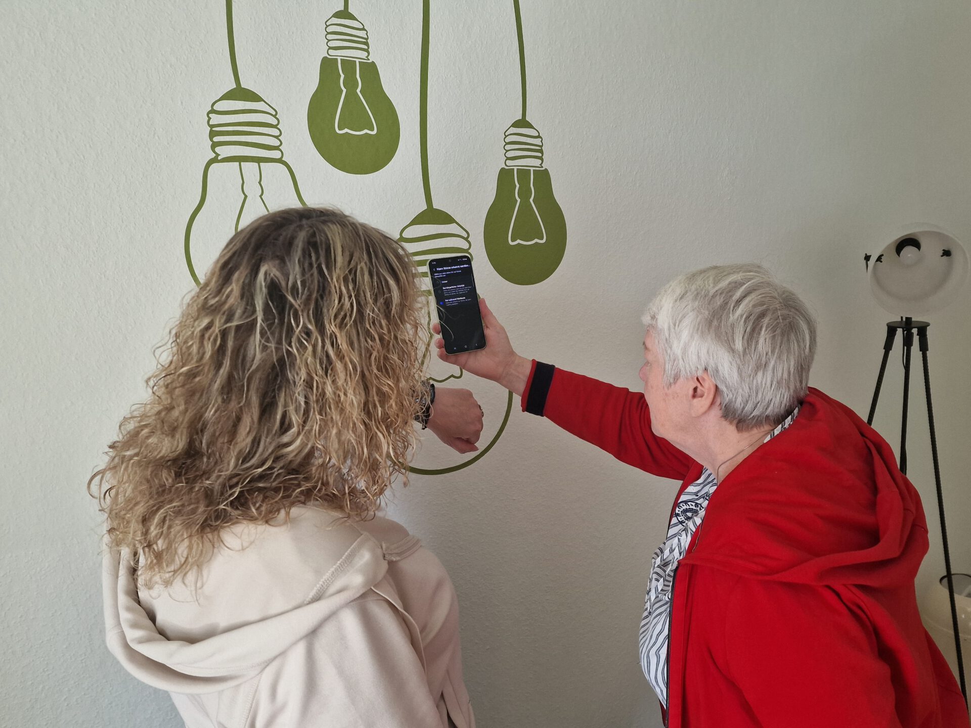 Zwei Frauen schauen auf eine Wand, auf der Glühbirnen gezeichnet sind. Die ältere Frau hat ein Smartphone in der Hand.