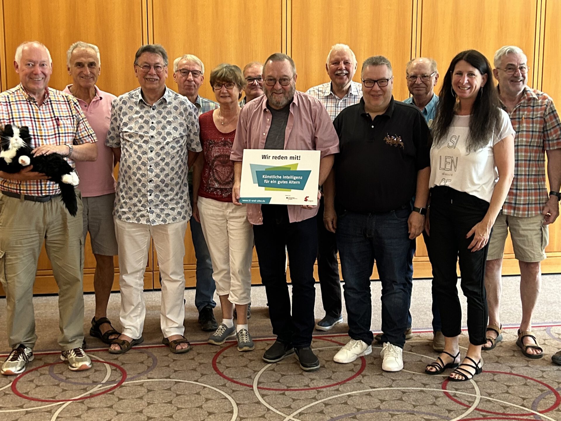 Eine Gruppe Personen lächelt. Eine Mann präsentiert ein Schild auf dem steht "Wir reden mit! KI für ein gutes Altern" Ein anderer Mann präsentiert eine Roboterkatze.