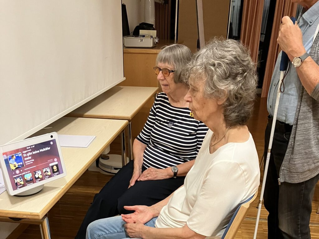 Zwei Frauen schauen sich einen smarten Lautsprecher mit Bildschirm an. Hinter Ihnen steht ein Mann mit Blindenlangstock.
