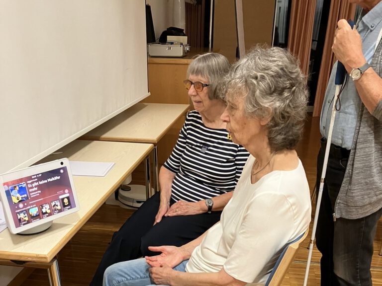 Zwei Frauen schauen sich einen smarten Lautsprecher mit Bildschirm an. Hinter Ihnen steht ein Mann mit Blindenlangstock.