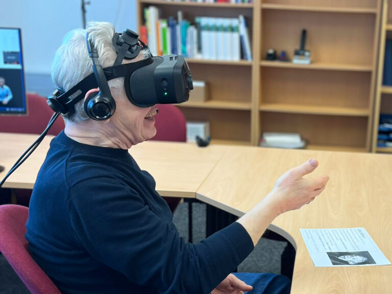 Ein älterer Herr mit VR_Brille, der lächelt und wie um jemanden die Hand zu reichen, seine Hand hebt