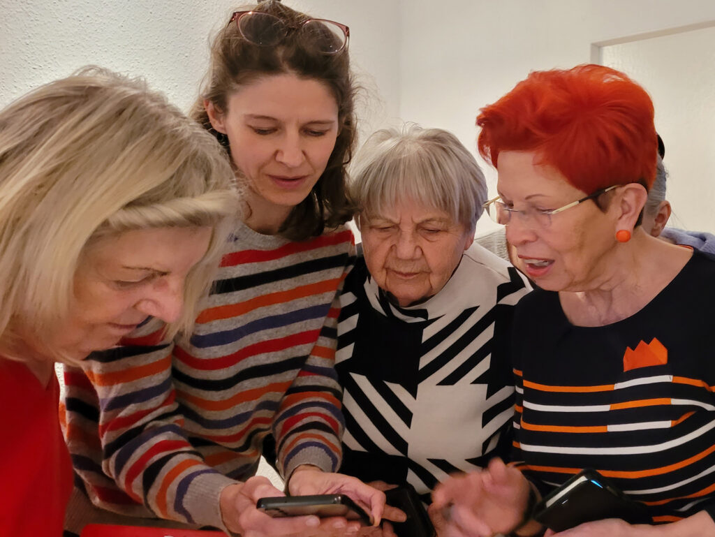 drei ältere und eine jüngere Frau schauen zusammen auf ein Smartphone