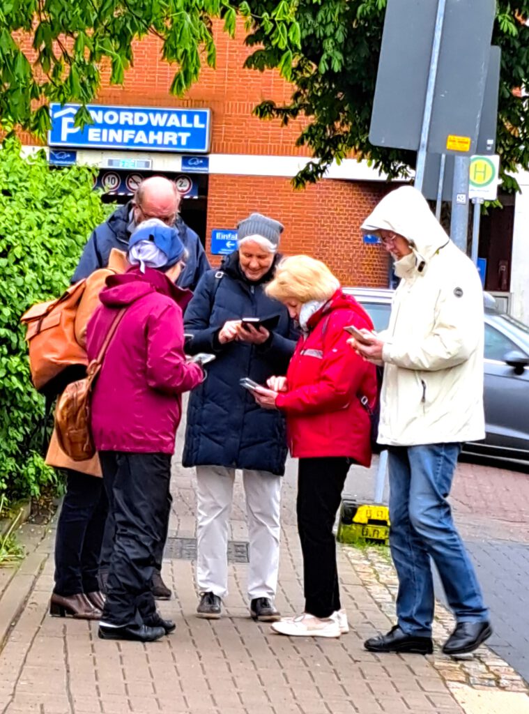 eine Gruppe älterer Menschen stehet vor einem Parkhaus. Alle schauen auf ihre Smartphones.