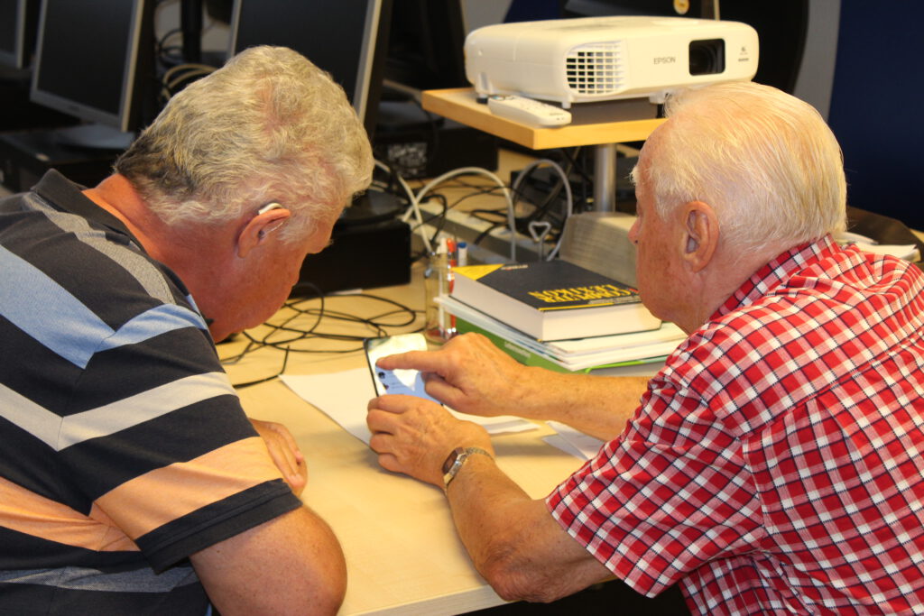zwei Männer, die auf ein Smartphone schauen. Der eine scheint dem anderen etwas zu zeigen.